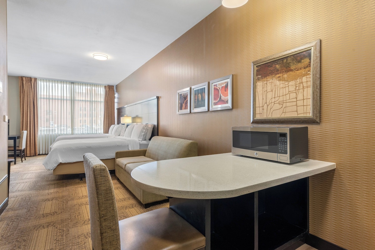A hotel room with a bed, a sofa, a dining table, a microwave, and a large window with curtains.