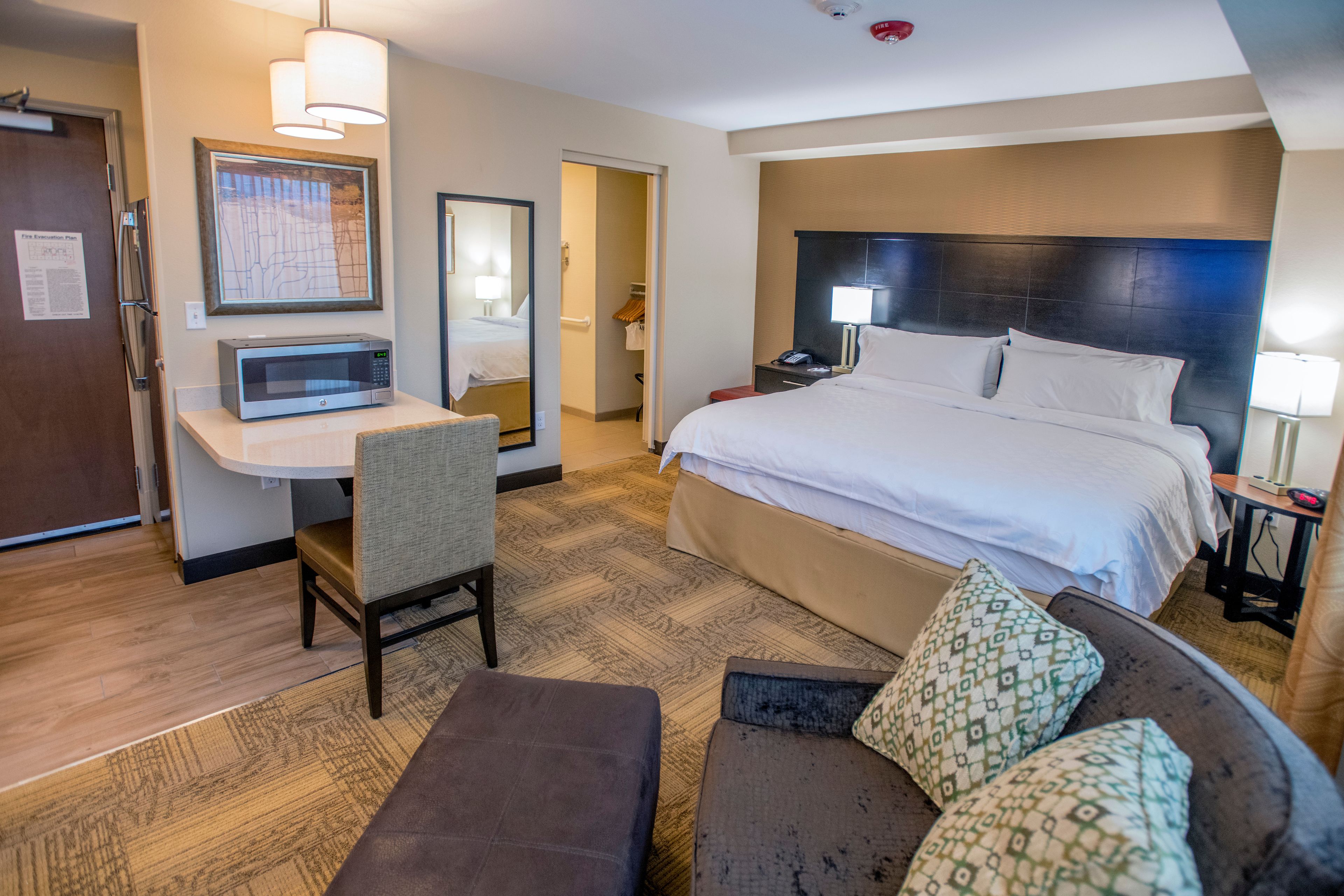 Habitación de hotel con cama, escritorio con microondas, una silla, un sofá y mesita de noche con lámpara.