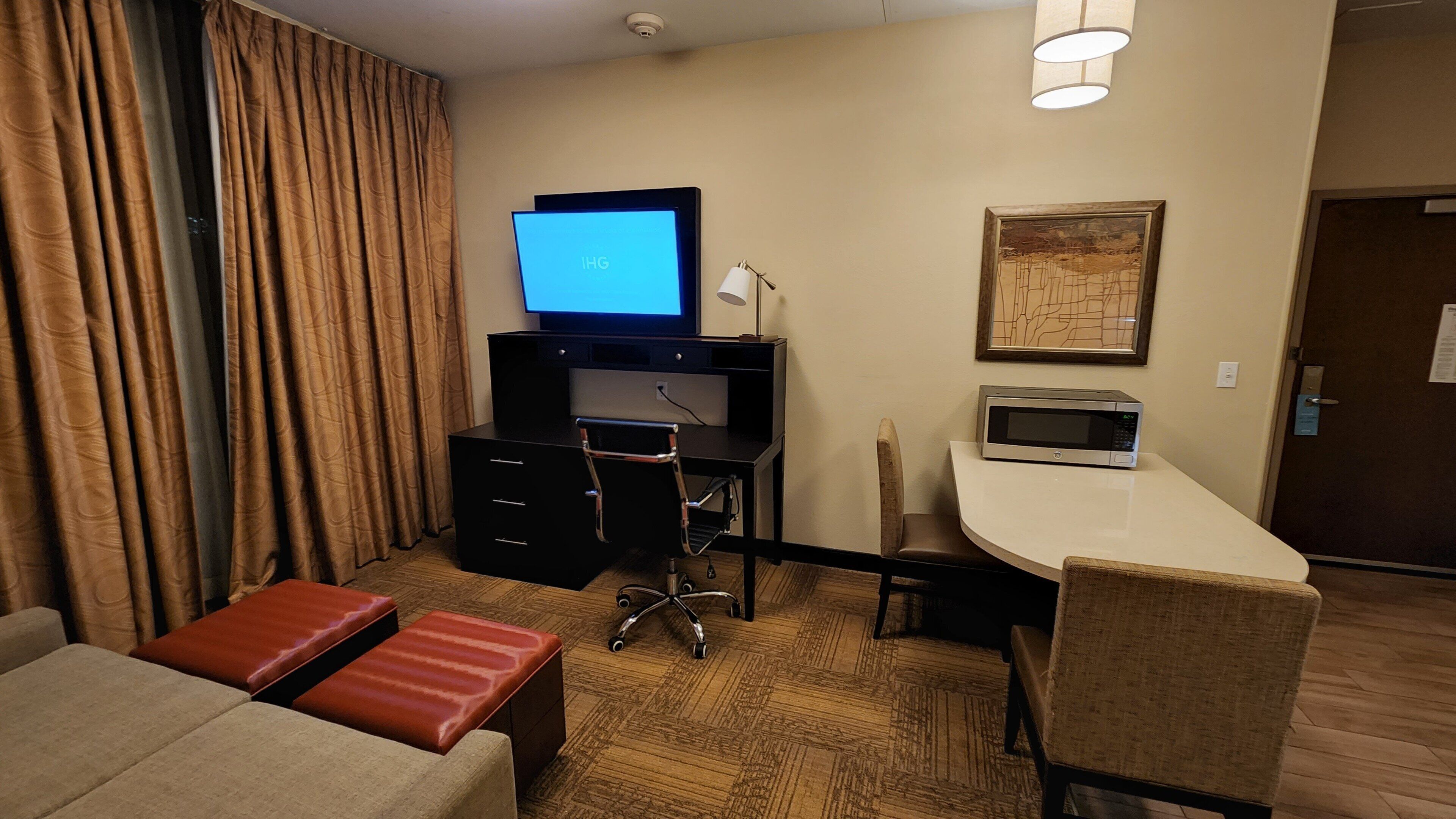 A hotel room with a desk, a chair, a couch, a red ottoman, a television, a microwave, and a framed picture on the wall.
