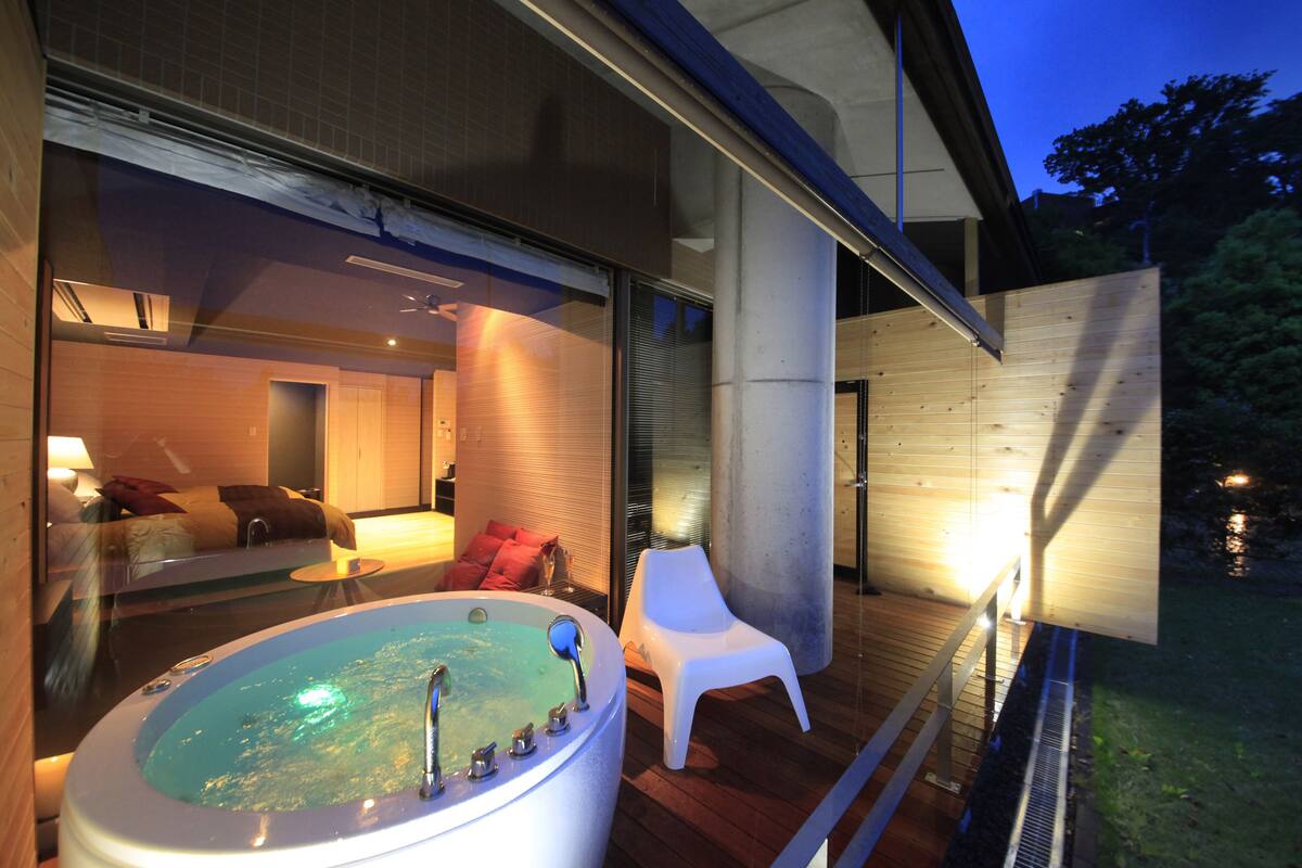 A modern outdoor hot tub area with a wooden deck, a white plastic chair, and a glass-enclosed room with a sofa and red pillows.