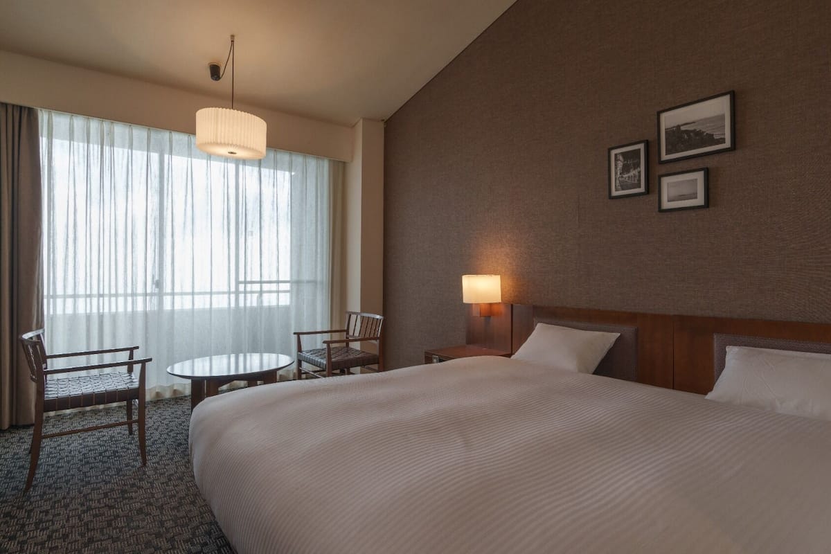 A hotel room with a large bed, a desk with a chair, a lamp, and a window with curtains.