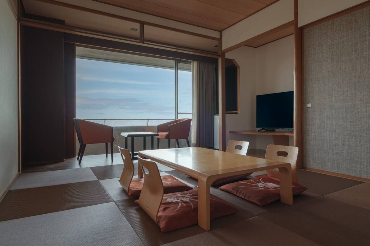 A room with a tatami mat floor, a wooden table with chairs, a television, and a view of the sea.