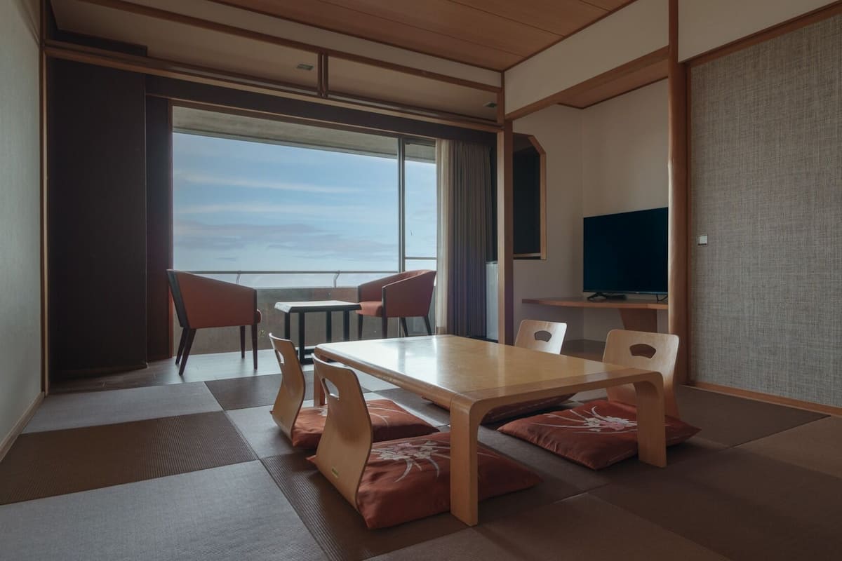 A room with a tatami mat floor, a wooden table with chairs, a television, and a view of the sea.