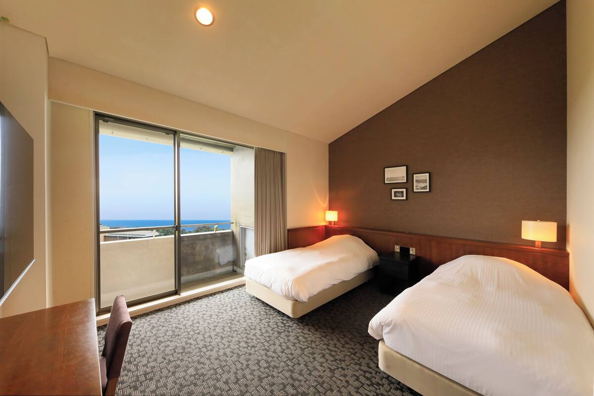 A hotel room with two beds, a large window with a view, a desk, and a chair.