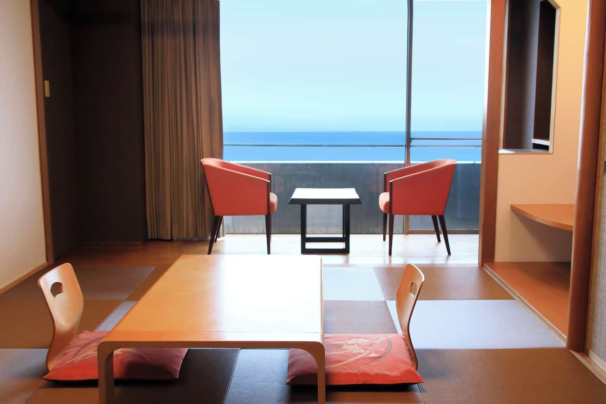 A room with tatami mats, a low table, two chairs, and a view of the sea.