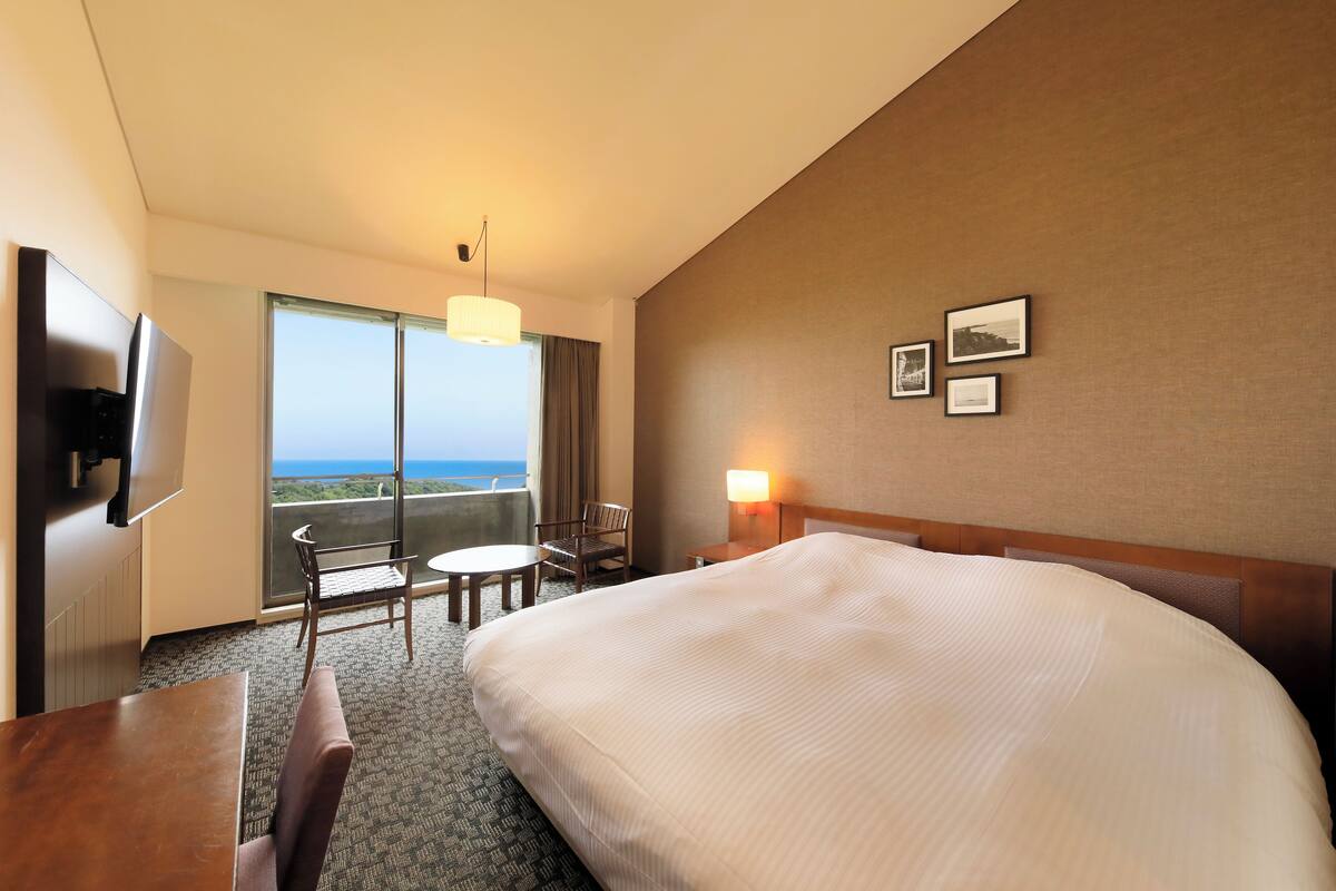 A hotel room with a large bed, a desk, a chair, a TV, a balcony with a view, and a wall-mounted lamp.