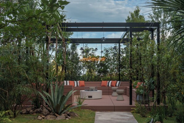Un espace extérieur moderne aménagé avec un coin salon, une pergola, un foyer et un panneau lumineux au néon.