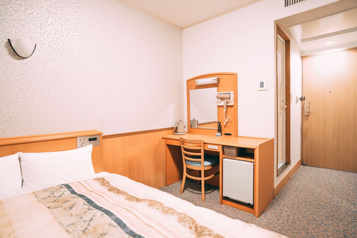 Habitación de hotel con cama, escritorio con silla, espejo, refrigerador pequeño y puerta.