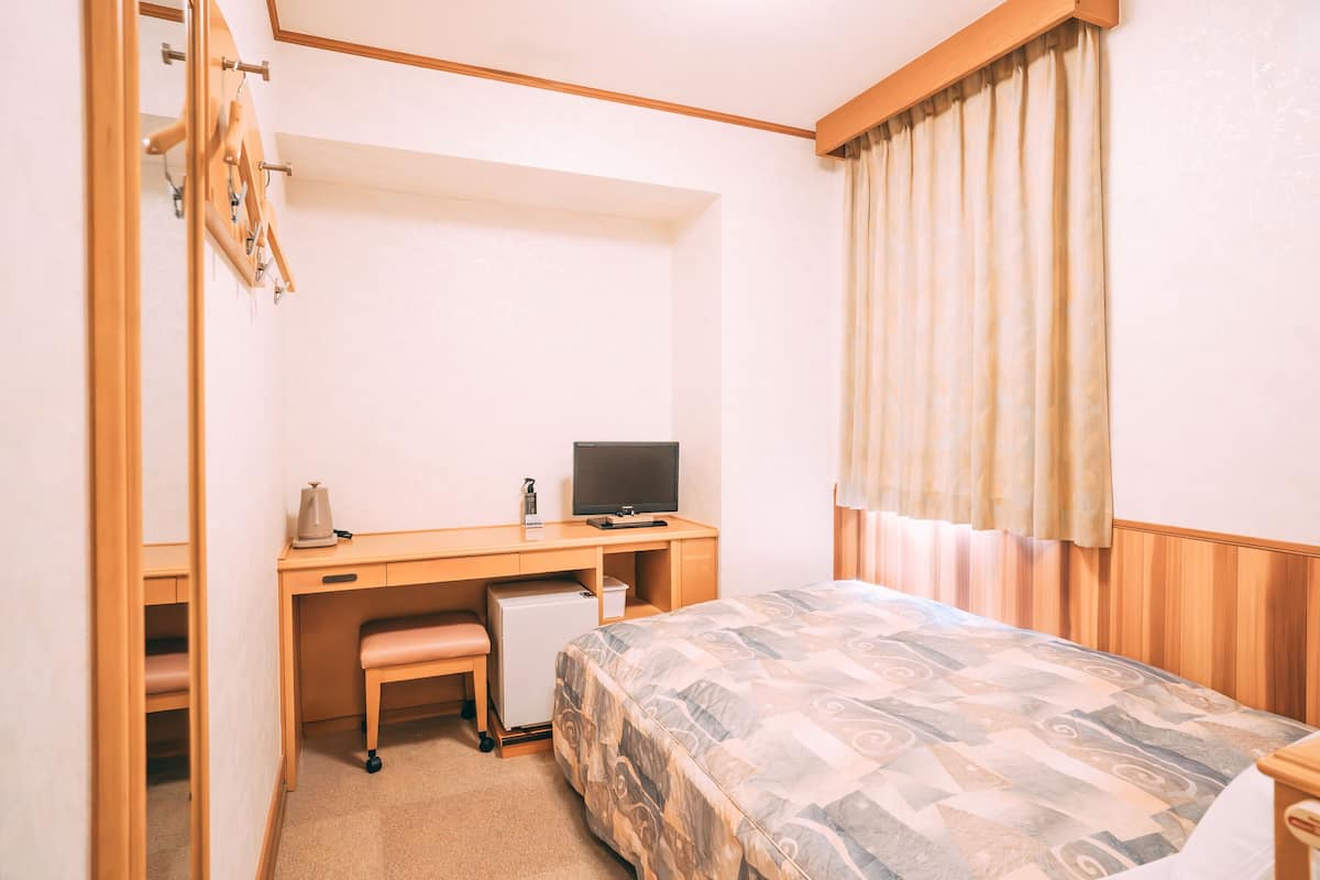 Habitación de hotel compacta con escritorio de madera, refrigerador pequeño, televisión, cama con ropa de cama estampada y ventana con cortinas.