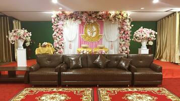 Espaço para eventos formais com um sofá de couro marrom, tapetes vermelhos e decorações florais.