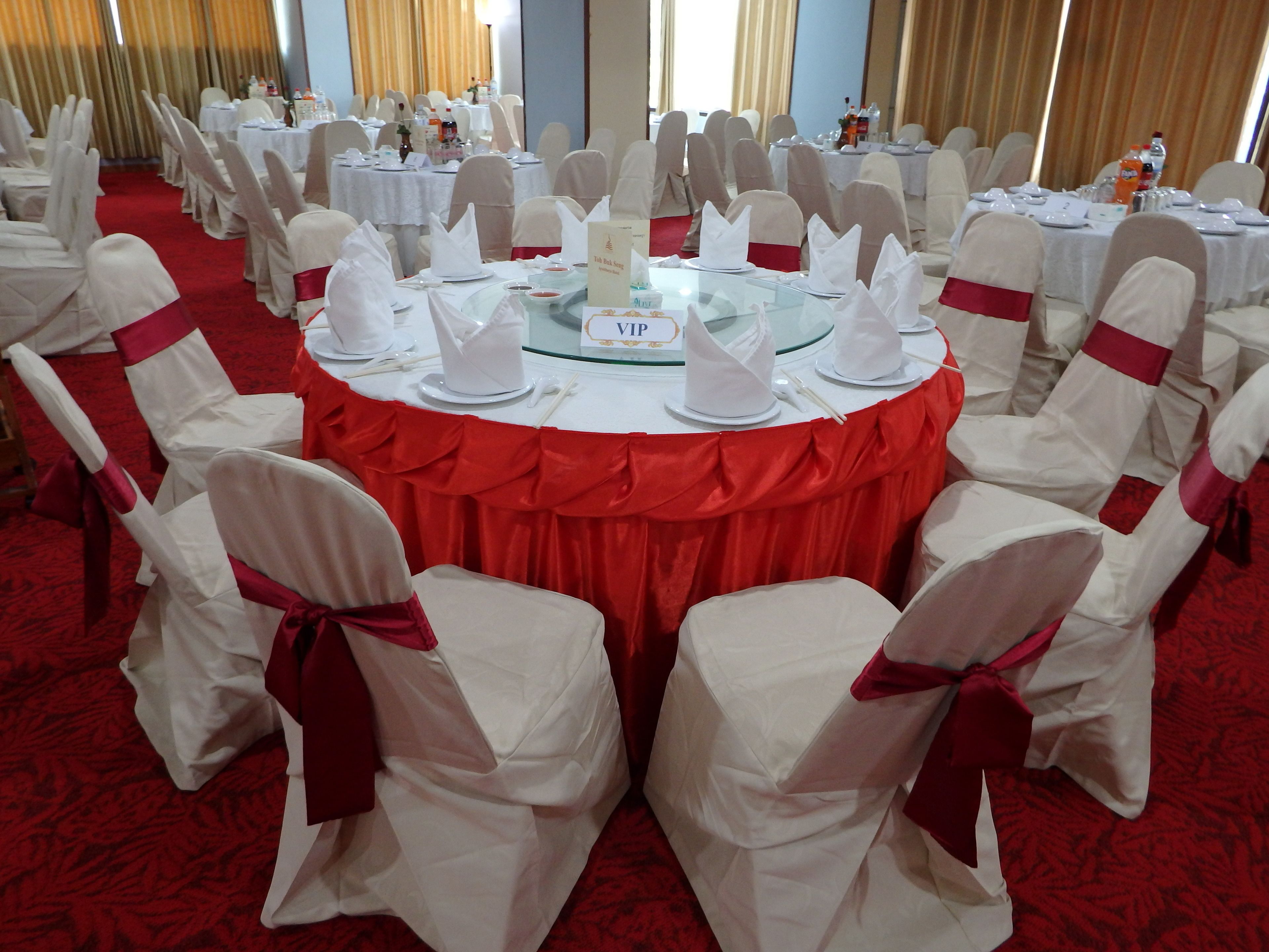 Un salón de banquetes con mesas redondas, sillas blancas con cintas rojas y una alfombra roja.