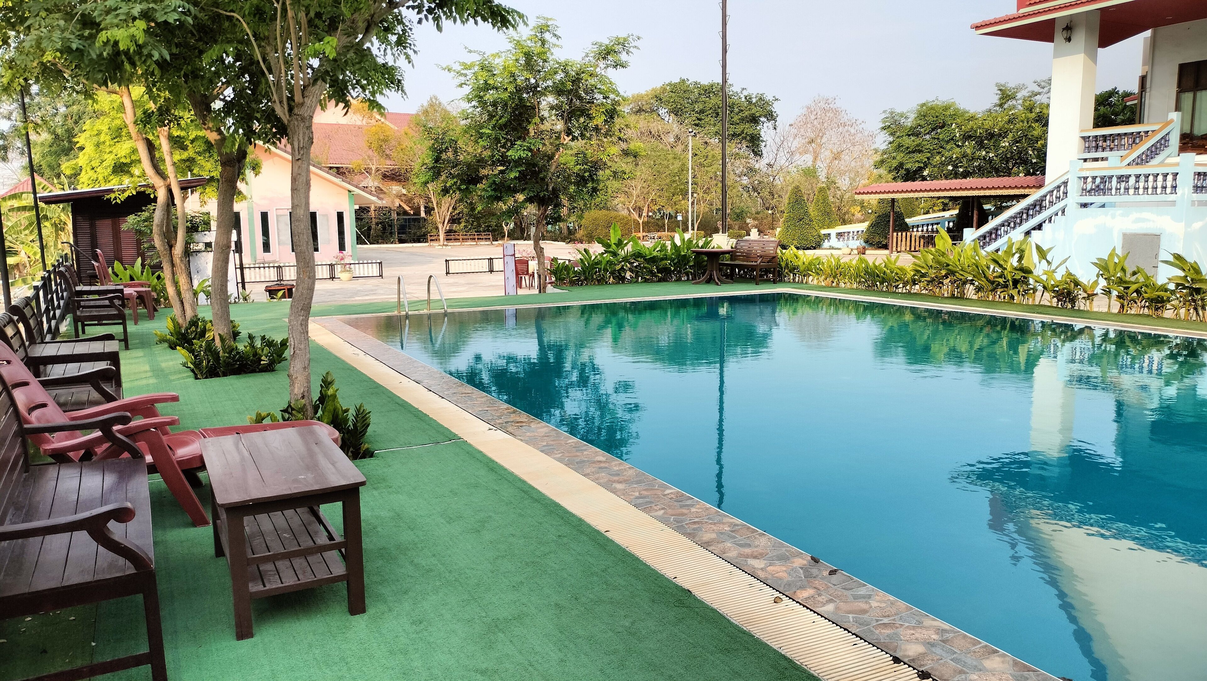 Una piscina con cubierta verde, sillas reclinables de madera y un edificio con techo rojo al fondo.