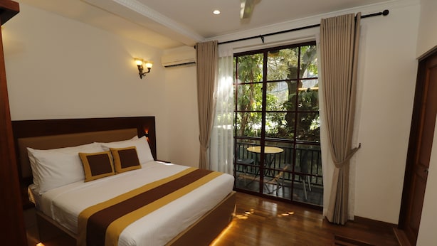 Une chambre d’hôtel avec un grand lit, un balcon doté d’une table et de chaises, et une vue sur la verdure environnante.