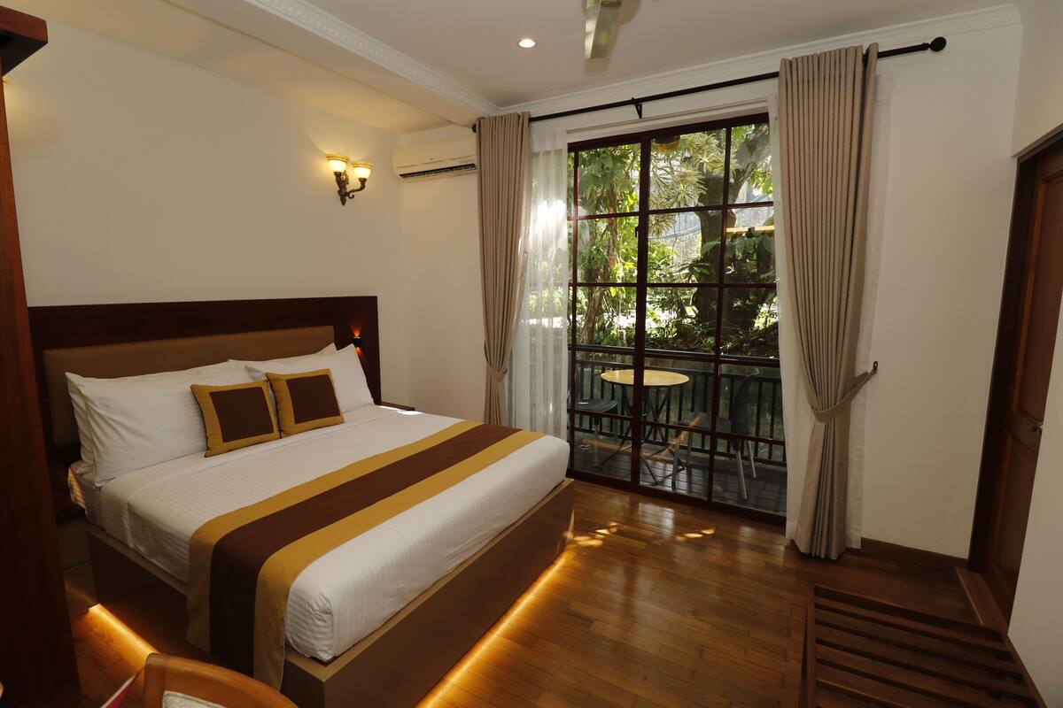A hotel room with a large bed, a balcony with a table and chairs, and a view of greenery outside.