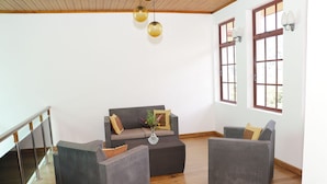 Un vaste espace de vie comprenant un canapé gris, deux fauteuils et une table basse. De grandes fenêtres aux montures marron et un plafond en bois complètent l’ensemble.