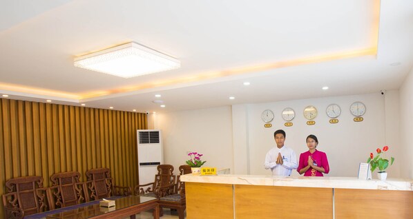 A hotel reception area with a reception desk, two staff members, a clock, and a potted plant.