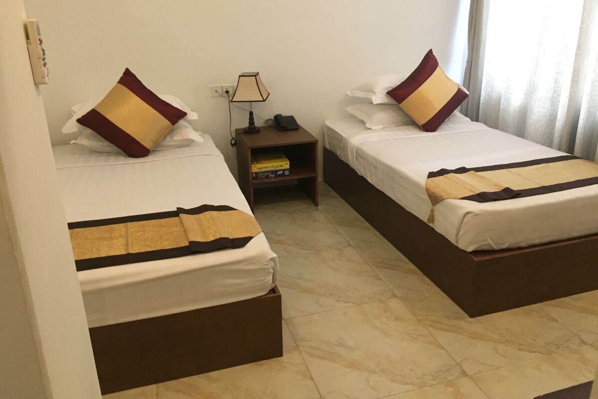 Une chambre d’hôtel avec deux lits simples, chacun équipé d’un oreiller et d’une couverture. Il y a une table de chevet avec une lampe et un livre. La chambre a un sol carrelé et une fenêtre avec des rideaux.