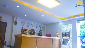 A hotel reception area with a wooden reception desk, multiple clocks on the wall, and a decorative flower arrangement. - Hotel Sunny Mandalay (Mandalay)