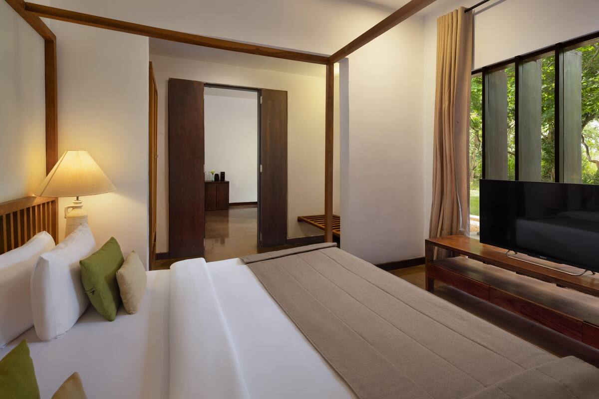 A modern hotel room with a large bed, a flat-screen TV, and a view of greenery through the window.