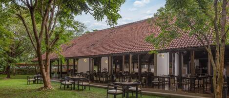 A restaurant with outdoor seating, covered by a roof, surrounded by trees and a well-maintained lawn.