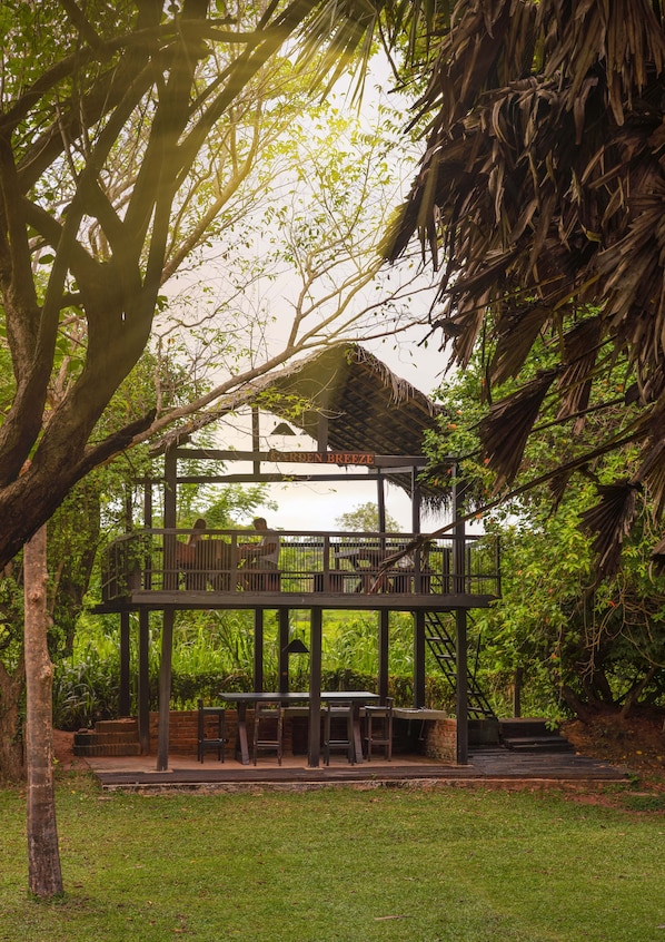 A wooden deck with tables and chairs, surrounded by lush greenery and trees.