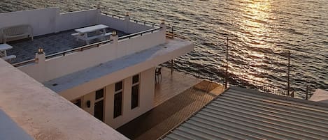 A seaside view with a sunset over the water, a deck with a table and chairs, and a building with windows.