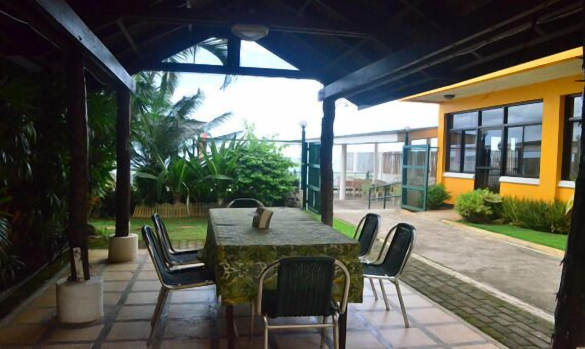 An outdoor patio with a table and chairs, surrounded by greenery and a building in the background.