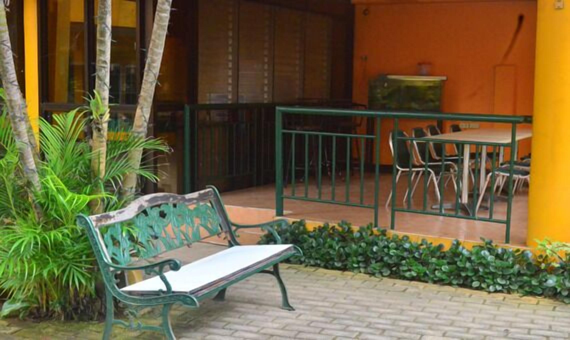 A bench and table set in a courtyard with a brick floor, surrounded by greenery and a yellow wall.