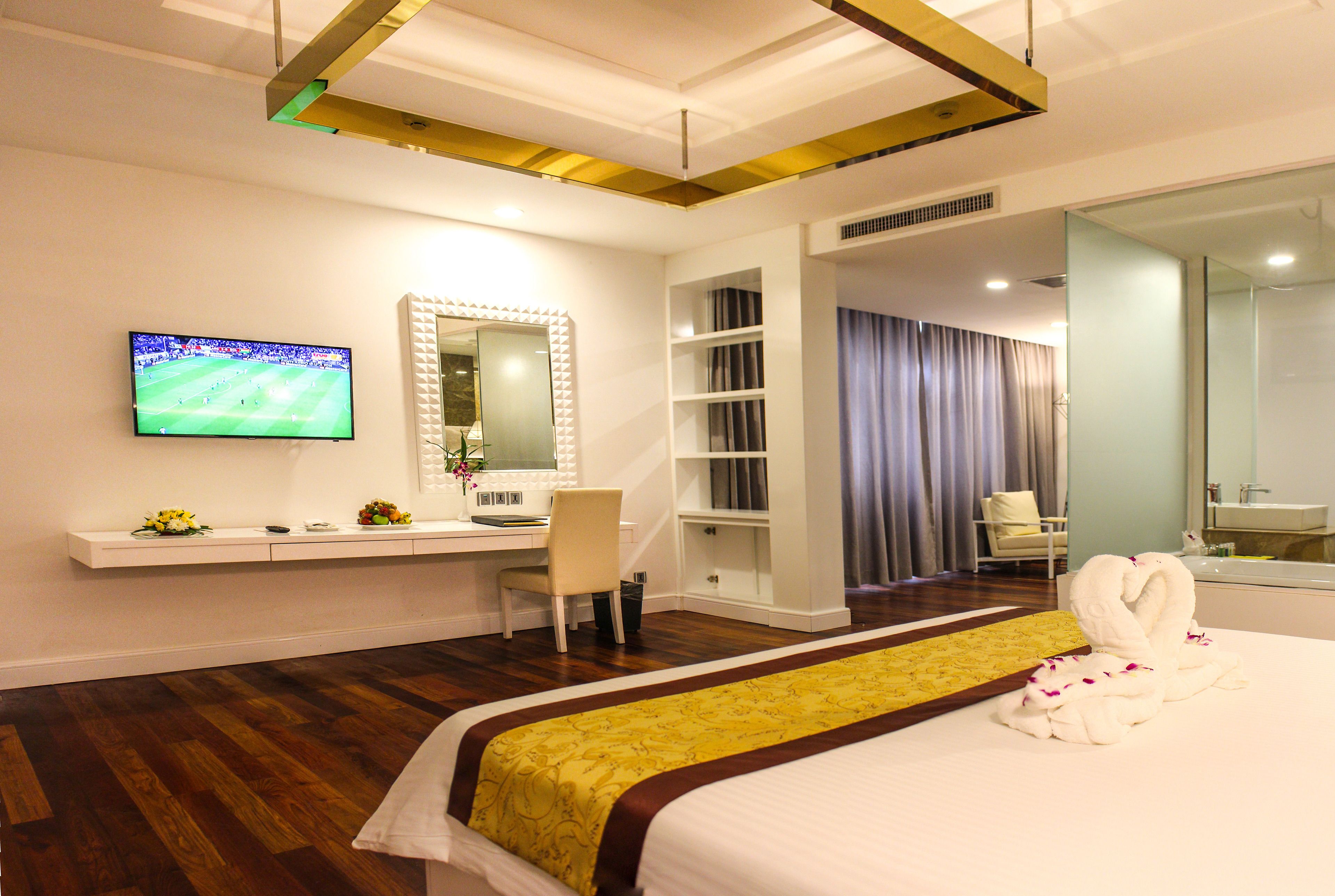 A hotel room with a bed, a desk with a computer, a TV, a mirror, and a bathroom visible through an open door.