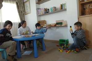A room with a blue table, children playing with toys, and bookshelves with books and decorative items. - Cimon Dolomites Hotel (Predazzo)