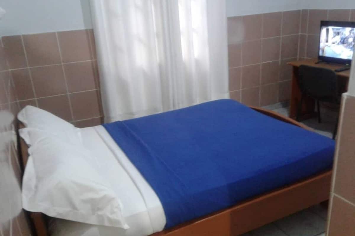 A single bed with a blue mattress and white pillows, a small desk with a chair, and a television on a stand.