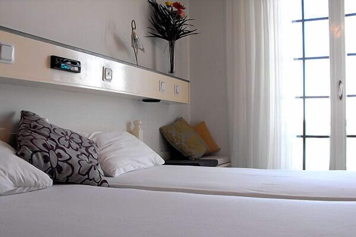 A hotel room with a bed, pillows, a bedside table, a vase with flowers, and a window with blinds.