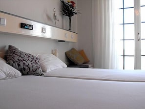 Hotellihuone, jossa on sänky, tyynyjä, yöpöytä, maljakko kukkineen ja ikkuna, jossa on sälekaihtimet.