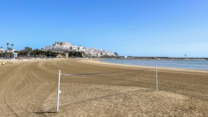 A beach with a sandy area and a volleyball net. In the background, there is a hill with buildings and a fort-like structure. - Apartamentos Voramar 3000 (Peniscola)