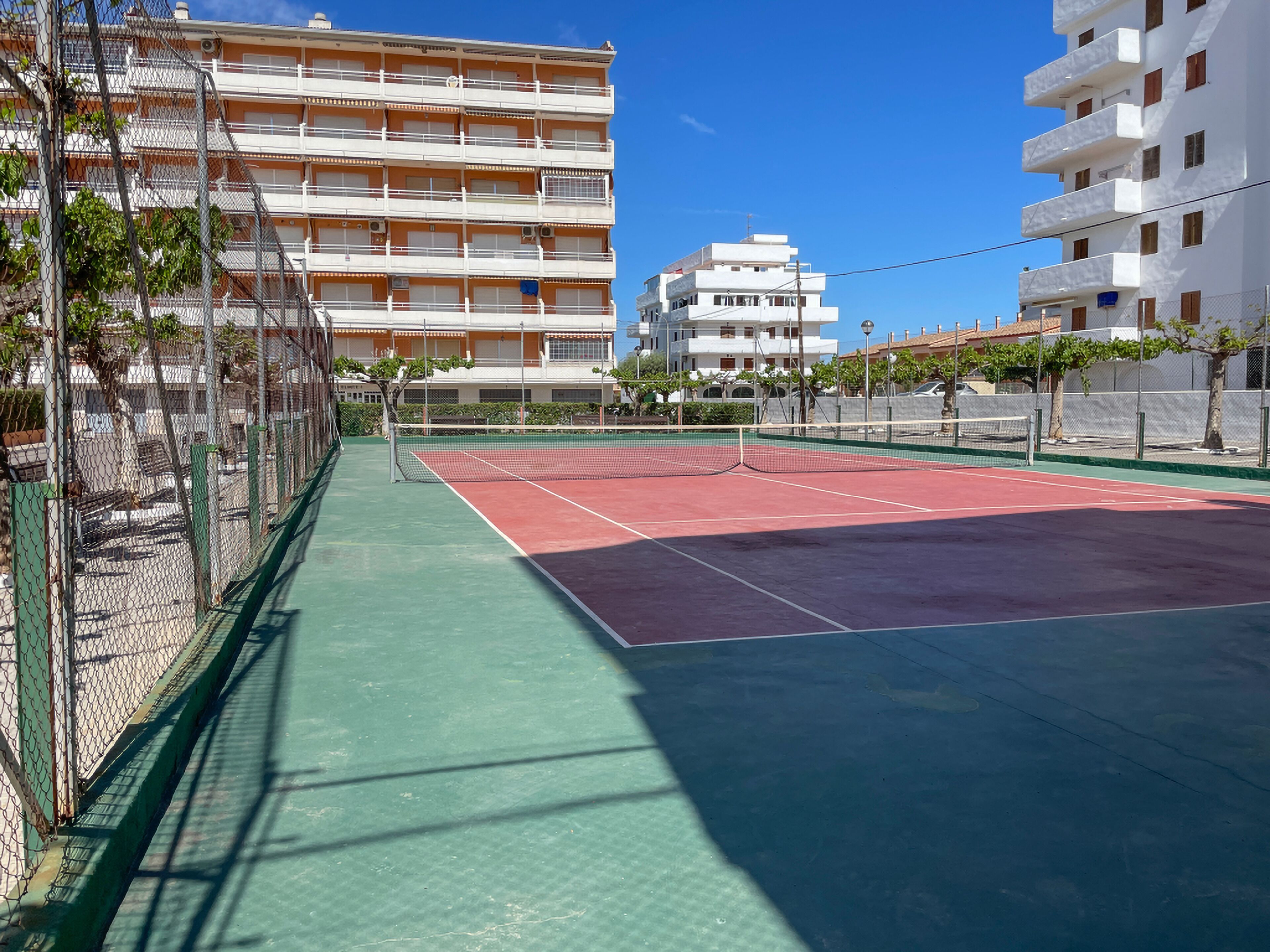 An outdoor tennis court with a green and red surface, surrounded by chain-link fencing and trees.