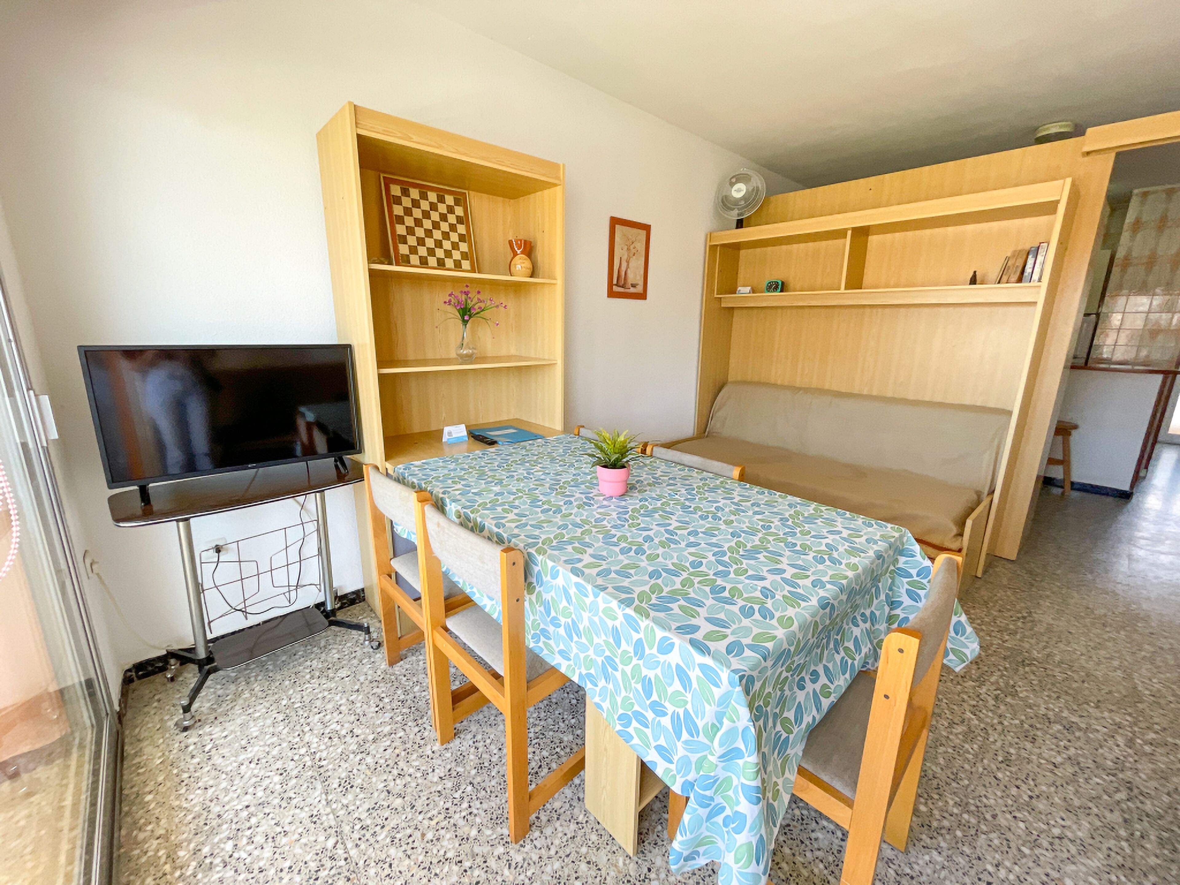 A compact living space with a TV, wooden furniture, a table with a patterned tablecloth, and a bookshelf.