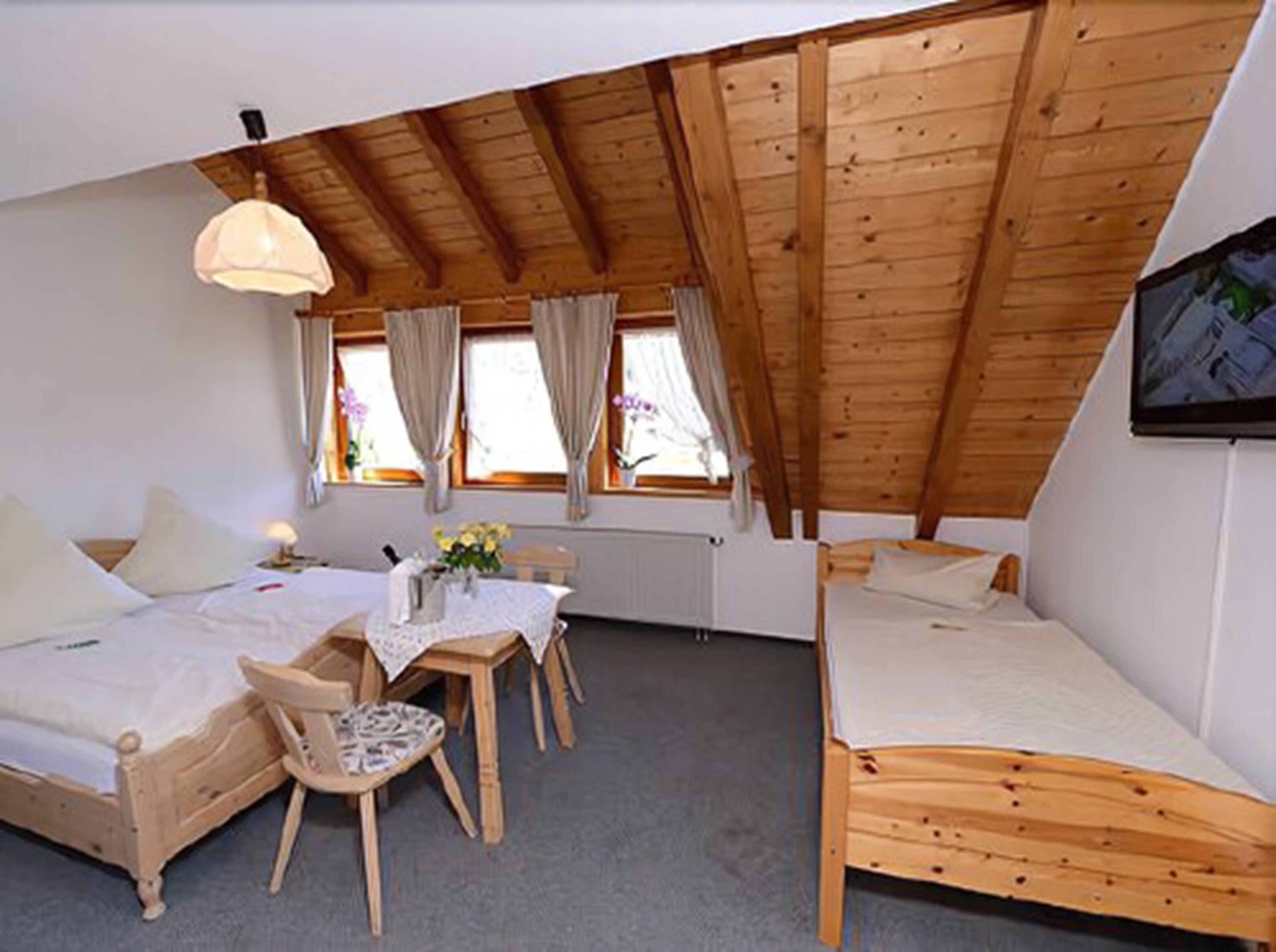 Ein gemütliches Zimmer mit Holzbalkendecke, zwei Betten, einem Tisch mit Stuhl und einem Fenster mit Vorhängen.