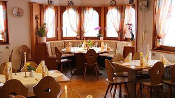 Un comedor con mesas y sillas de madera, una mesa redonda en el centro y ventanas grandes con cortinas.