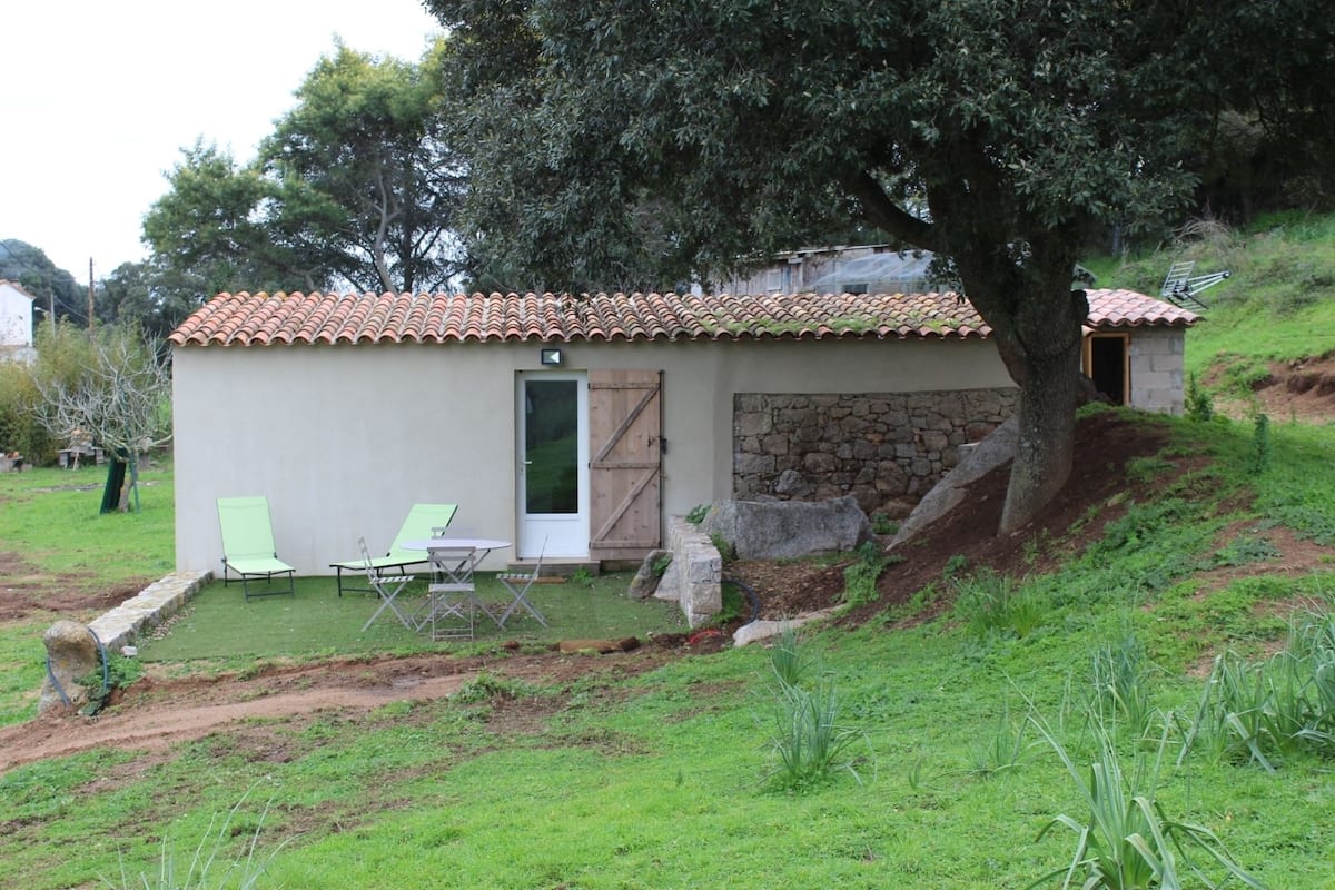 A small, single-story building with a tiled roof, a stone wall, and a door. There are two green chairs and a table outside.