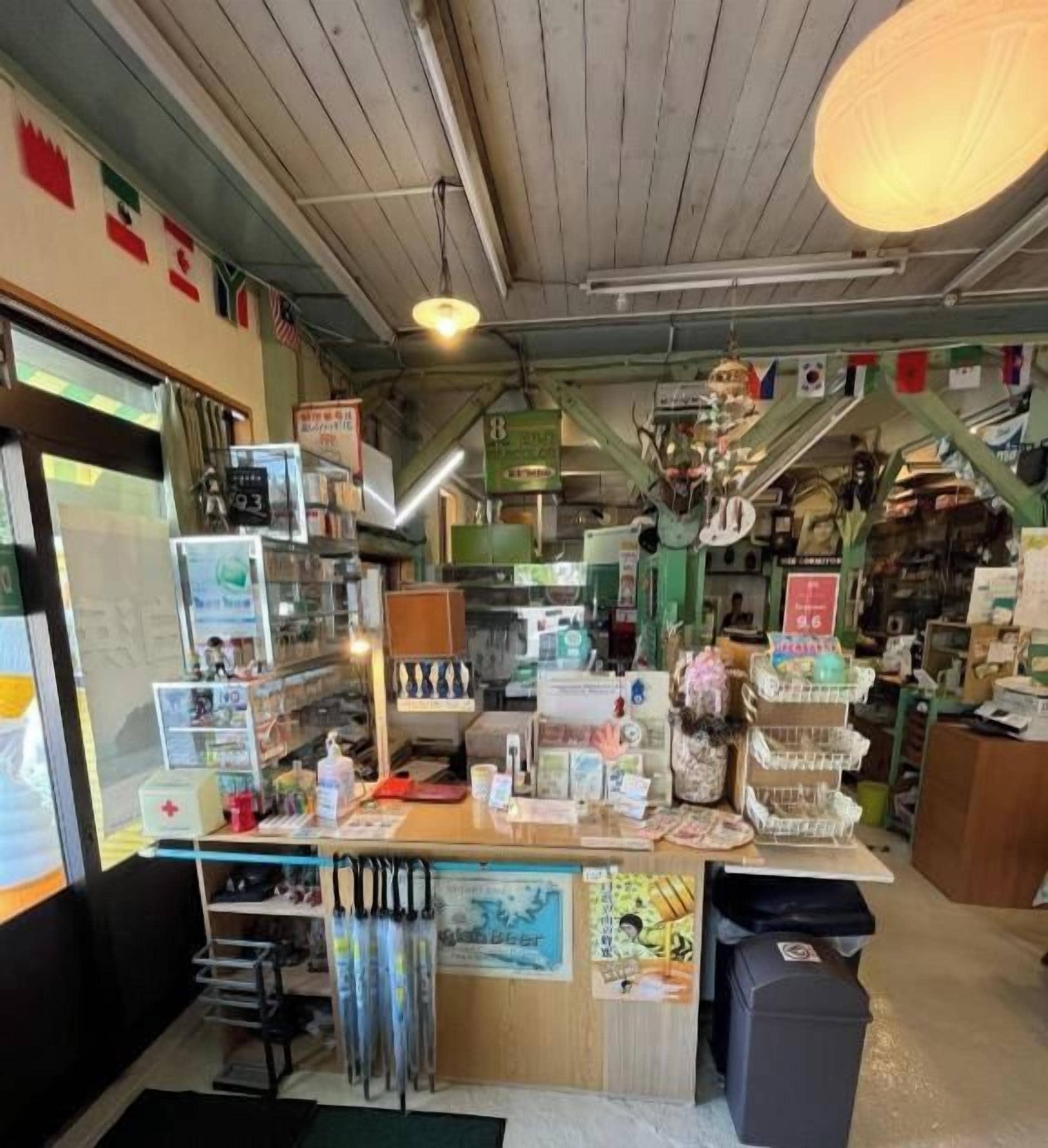 A small, cluttered shop with various items on shelves and counters, including a cash register and a trash bin.