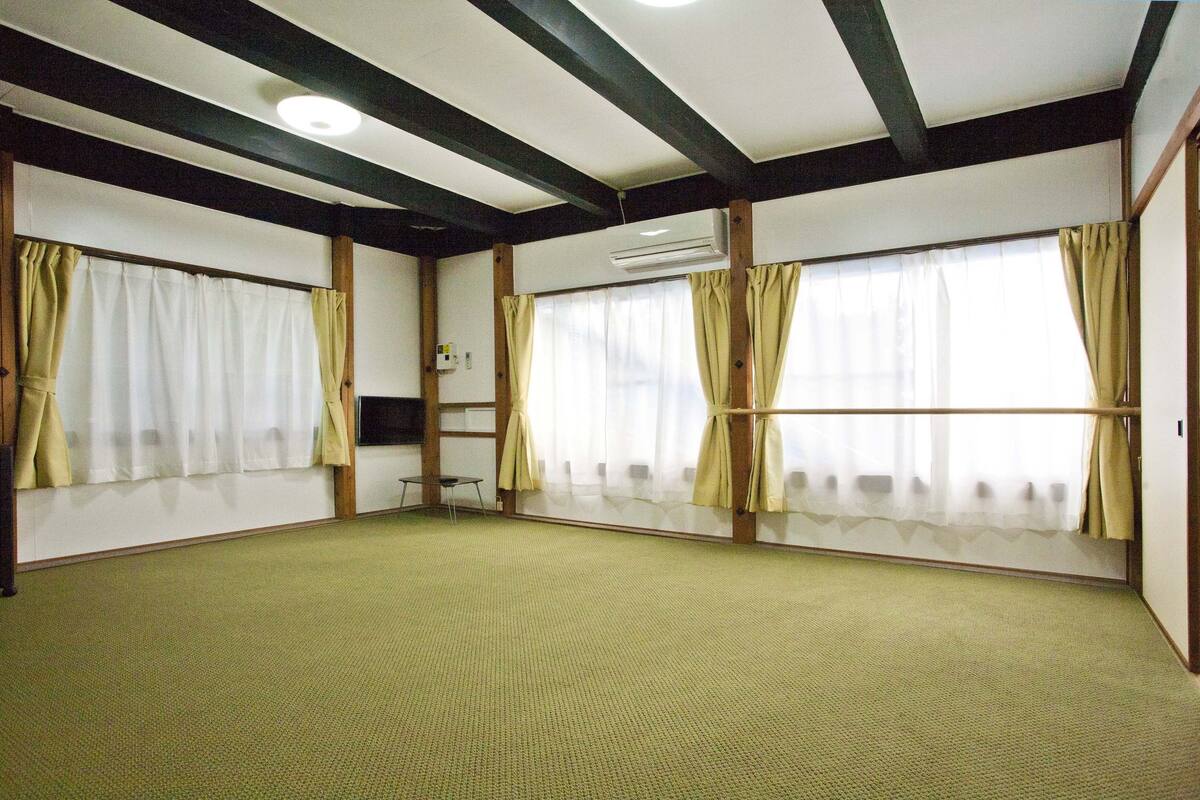 Quarto com carpete verde, vigas de madeira no teto e grandes janelas com cortinas.