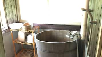 A rustic bathroom with a large wooden tub, a metal shelf with a bowl, and a window with blinds.
