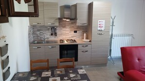 Cocina pequeña con estufa a gas, pileta y muebles de madera. Se observa una silla roja y una mesa con colcha.