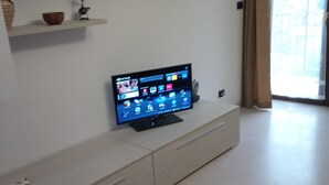 Una sala de estar con un televisor de pantalla plana sobre un mueble bajo, un estante blanco con adornos y un ventanal con cortinas.