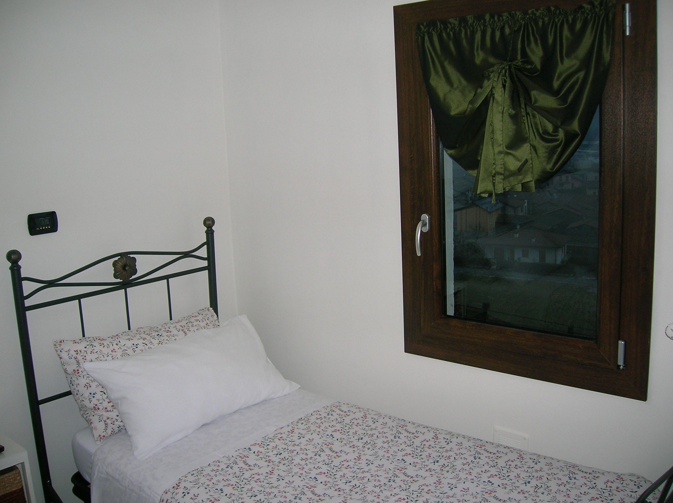 Quarto pequeno com uma cama de solteiro, cabeceira de madeira, janela com cortina verde e termostato fixado na parede.