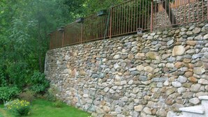 Un muro de piedra con barandilla metálica, rodeado de follaje y una zona ajardinada con escalones que conducen hacia él.