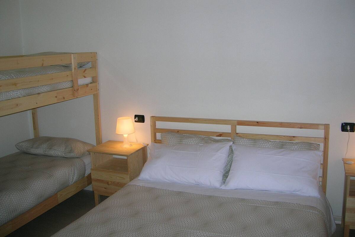 A bunk bed with a wooden frame and a single bed with white bedding. There is a nightstand with a lamp and a power outlet on the wall.