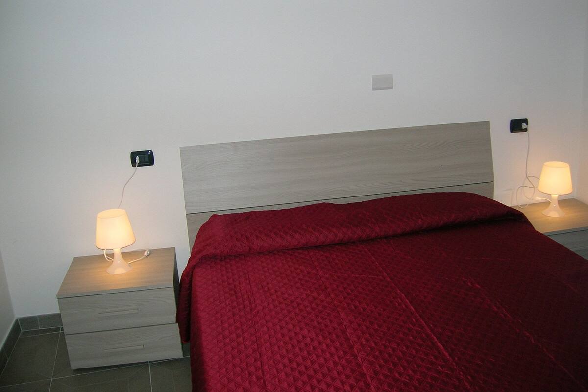 A bed with a red quilt, two bedside tables with lamps, and a wall socket.