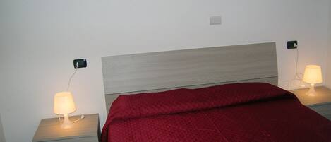 Una cama con colcha roja, dos mesitas de noche con lámparas y un enchufe de pared.