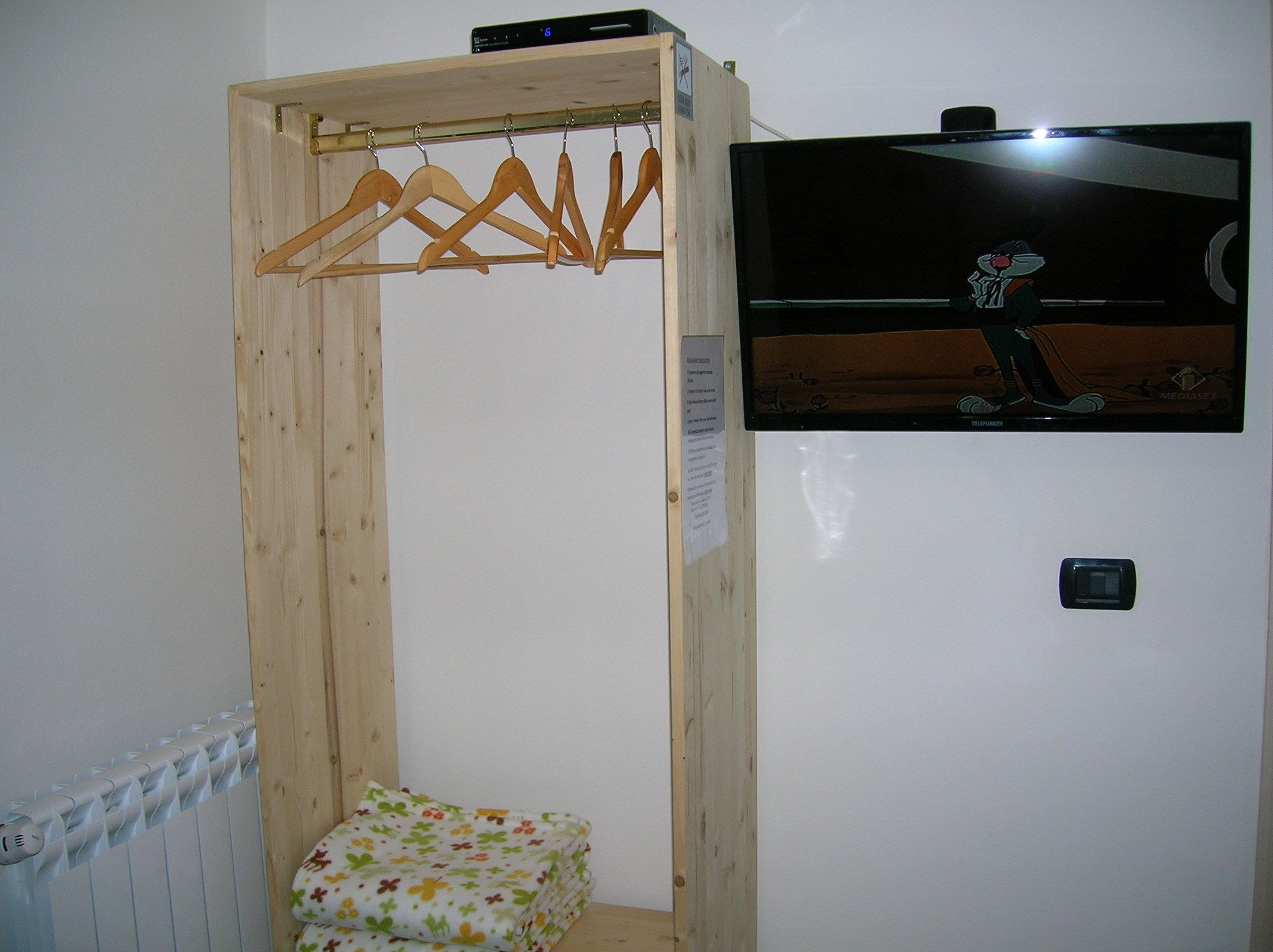 A wooden wardrobe with a hanging rack, a small bed with a floral bedspread, and a television mounted on the wall.